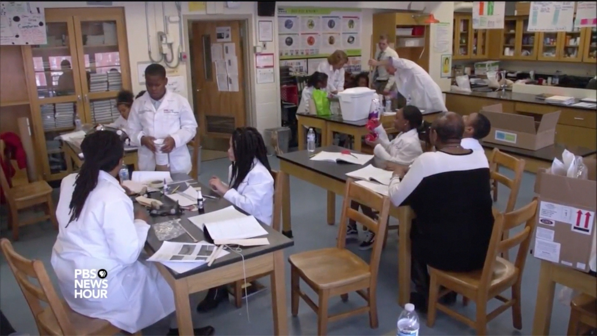 CURE Scholars at Franklin Square Elementary/Middle School work on science projects as shown in a video report by PBS NewsHour. Core team members for the CURE Scholars Program include Brian Sturdivant, MSW, far left, and Executive Director Robin Saunders, EdD, MS, mentoring a student at a table, far right.  