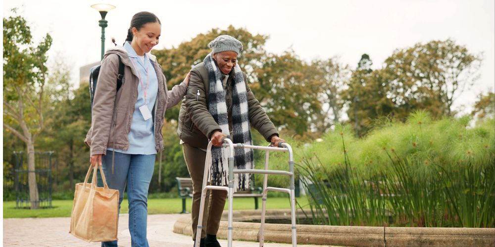 Stock image of an older woman using a walker, accompanied by a caregiver who has a hand rested on her shoulder as they walk.