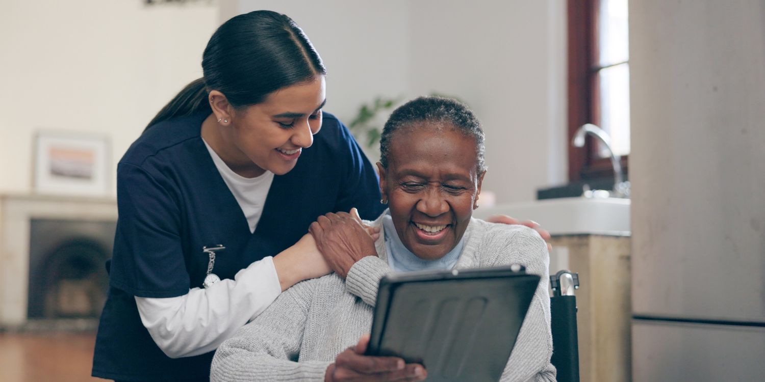 Stock image of an older patient looking at an electronic device, accompanied by a caregiver who has a hand on her shoulder. 