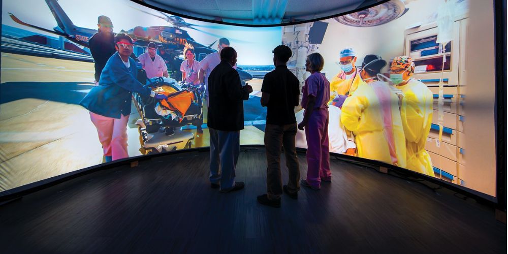 Three individuals stand in the center of a room surrounded by a floor-to-ceiling curved screen that is displaying images of trauma care.