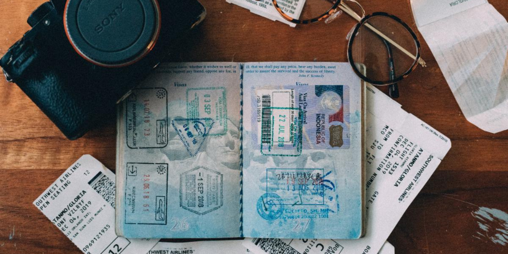 A passport open on a table along with a camera, plane tickets, and glasses.