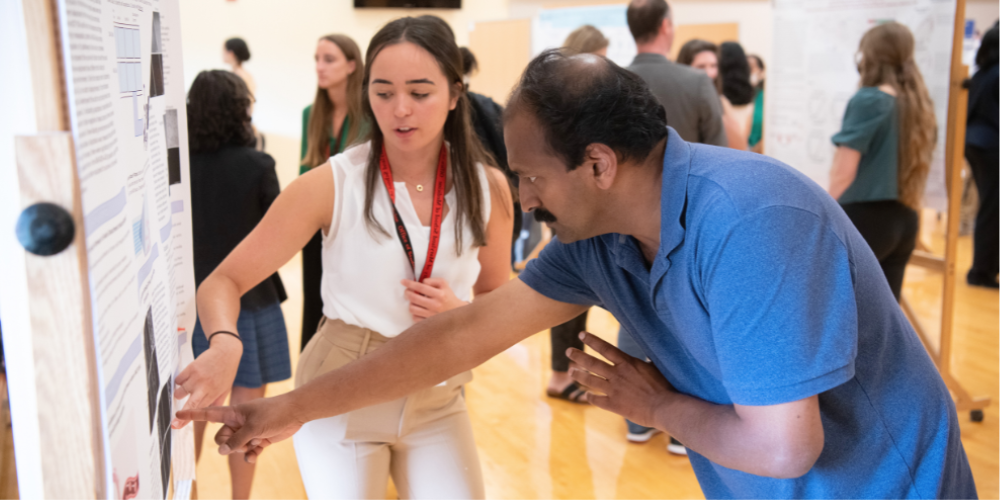 A student and faculty member point to something on the student's poster during a poster session.