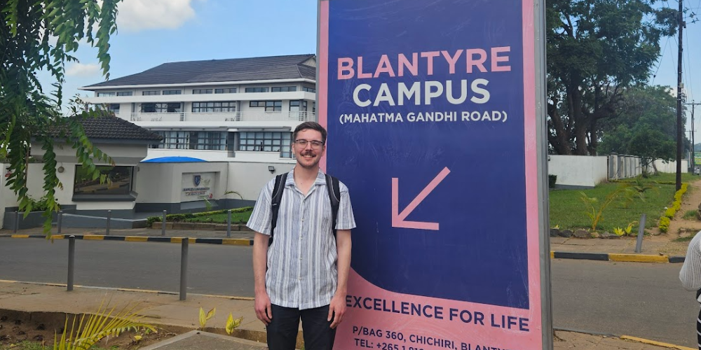 A student stands outside beside a sign for Kamuzu University's Blantyre Campus