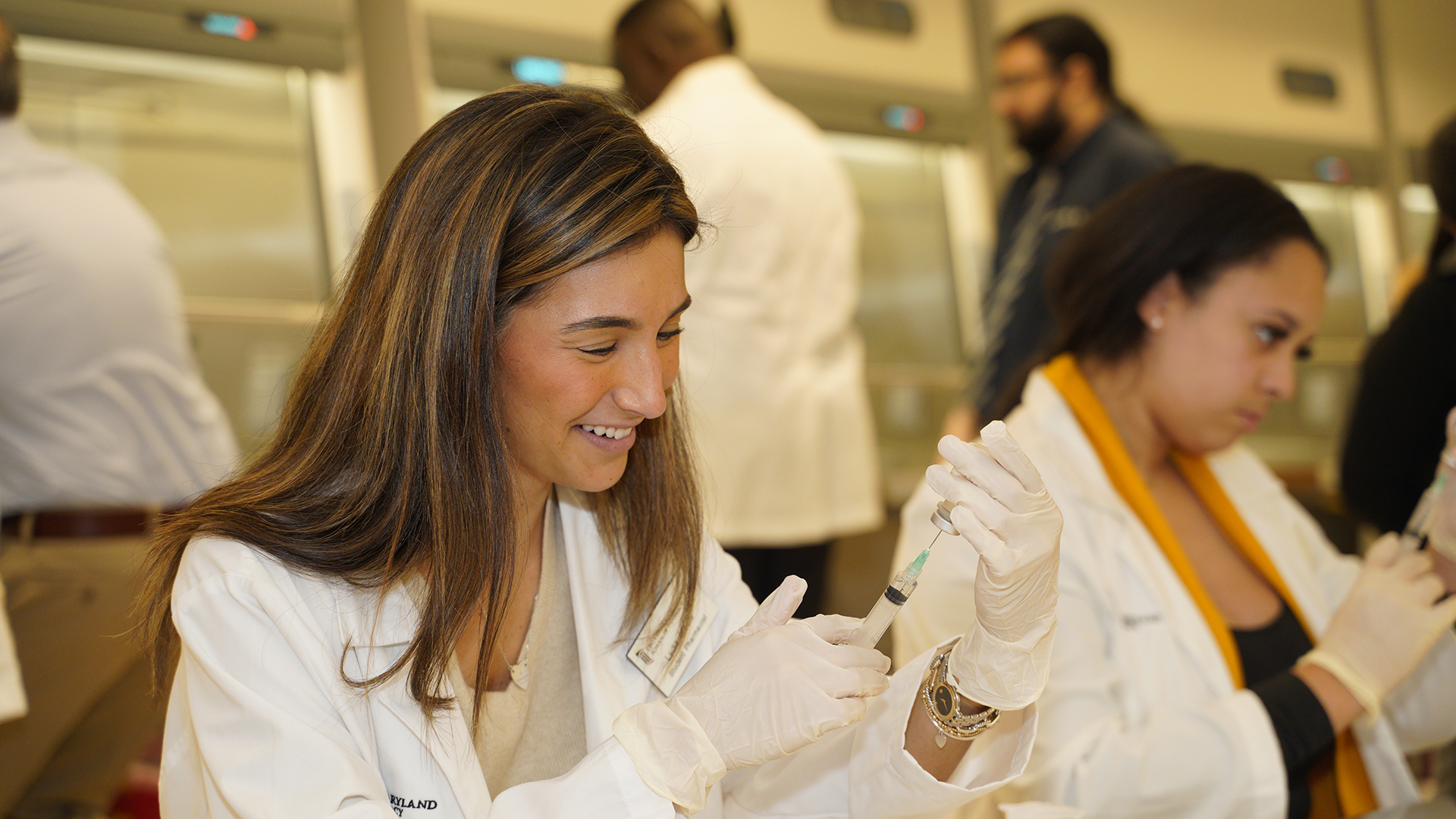A pharmacy student drawing a medication into a needle, in a lab with other students around her.