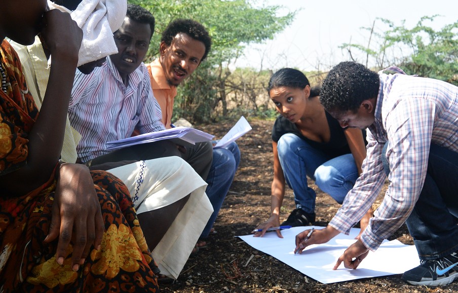 Man and woman squat to look at a large paper, while others look on.