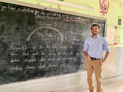 Man in blue shirt and khaki pants in front of school blackboard with writing on it