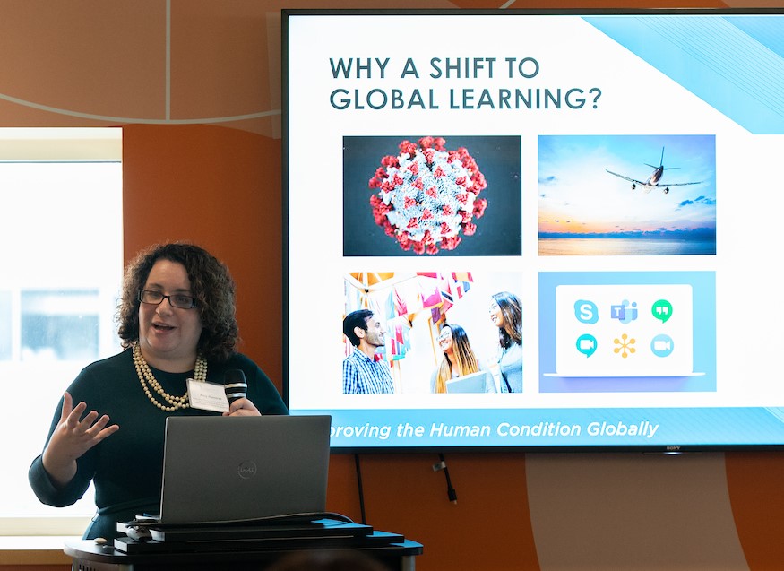 A woman speaks at a podium in front of a PowerPoint presentation on-screen