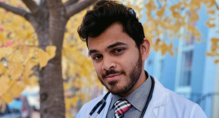 Man wearing white medical jacket and stethoscope posing outside with a fall-colored tree behind him