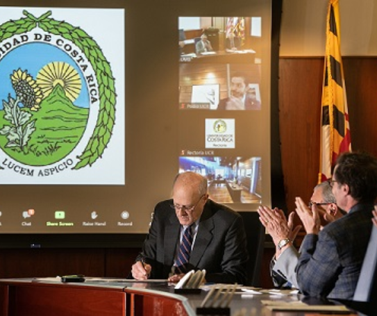 President Bruce Jarrell signs an MOU while two others look on with a video screen behind him