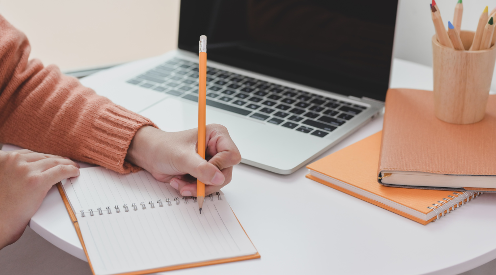 Hand holding a pencil writing in a notebook with a laptop also on the table