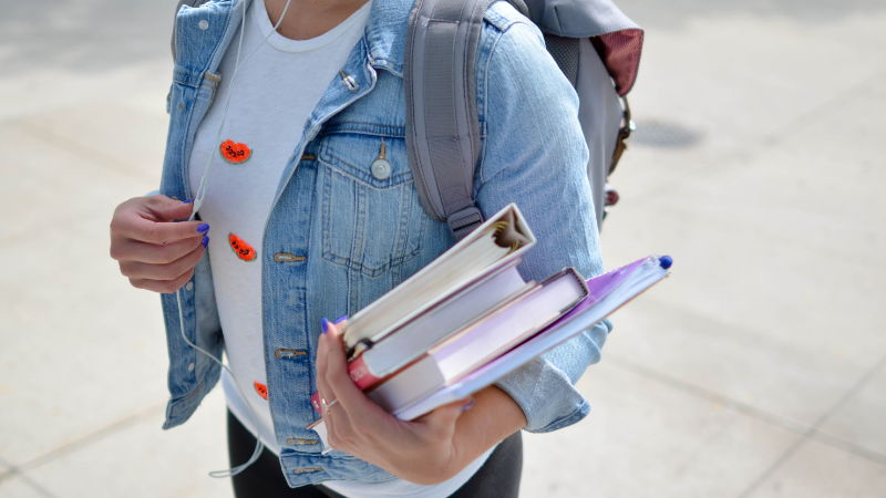 Female student in jean jacket carries school books in her left arm