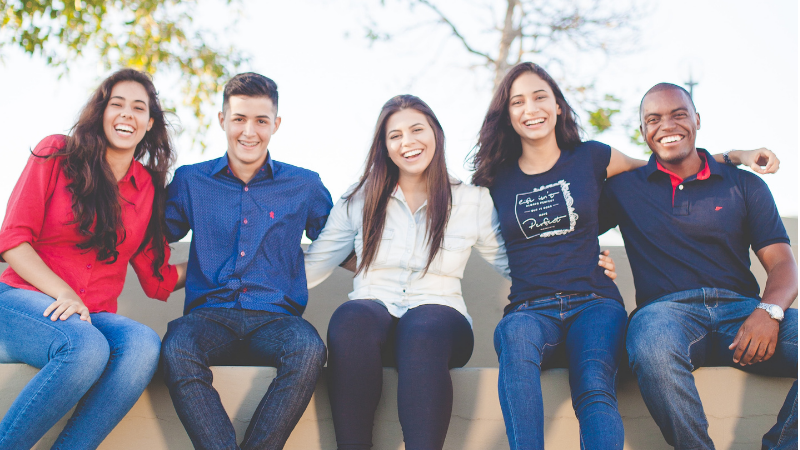 Diverse group of five students sit on low wall with their arms around each others shoulders