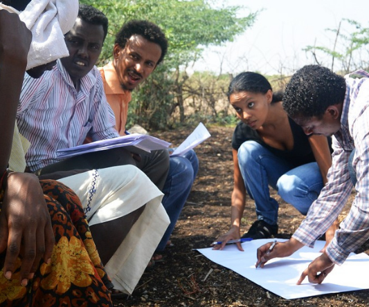 A man and woman squat to look at a large paper while others look on