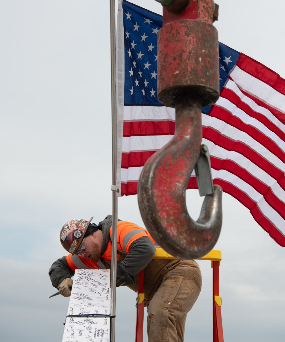 Construction worker in safety gear marking a metal beam with an American flag and large crane hook in the foreground.