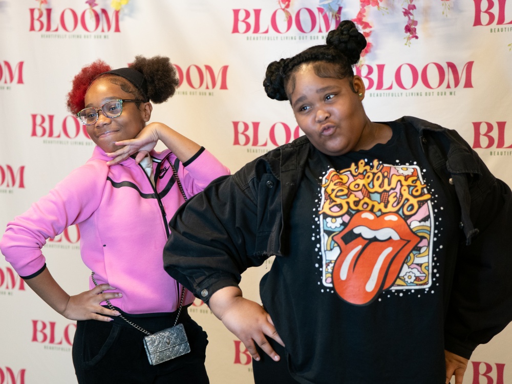 Two smiling girls pose playfully in front of a “BLOOM: Beautifully Living Out Our Me” step-and-repeat backdrop, striking confident, fun stances. One wears a pink hoodie and glasses with her hand under her chin; the other wears a black jacket and a Rolling Stones T-shirt, puckering her lips and posing with attitude.