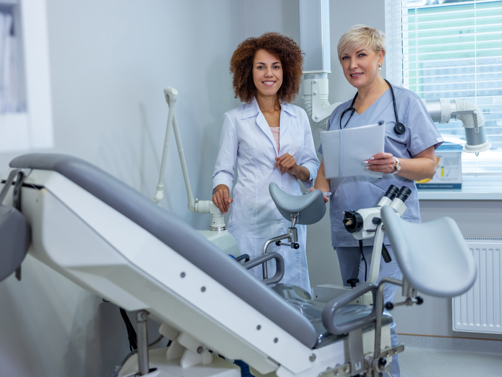 Two health care professionals stand beside a gynecological exam table and medical equipment in a clinical setting.
