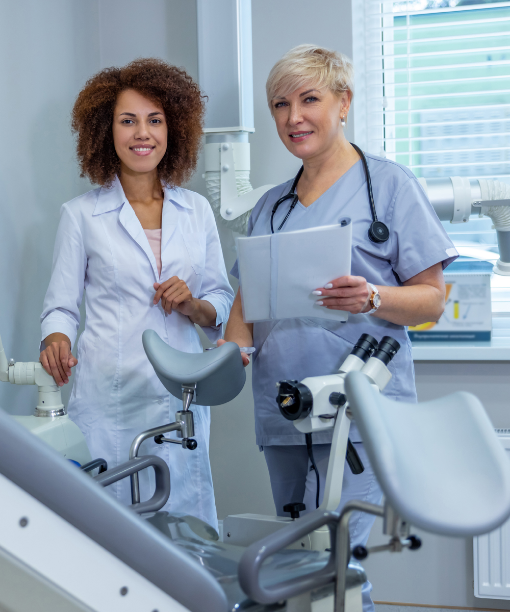 Two health care professionals stand beside a gynecological exam table and medical equipment in a clinical setting.