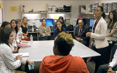 Elsie Stines, MS, CPNP, stands amid students and faculty including President Jay A. Perman, MD, who sits to her right, during the interdisciplinary President's Clinic.