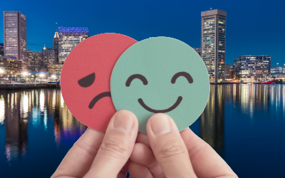 A hand holding two circle paper cutouts, a green smiling face and a red frowning face against a backdrop of Baltimore's Inner Harbor at night