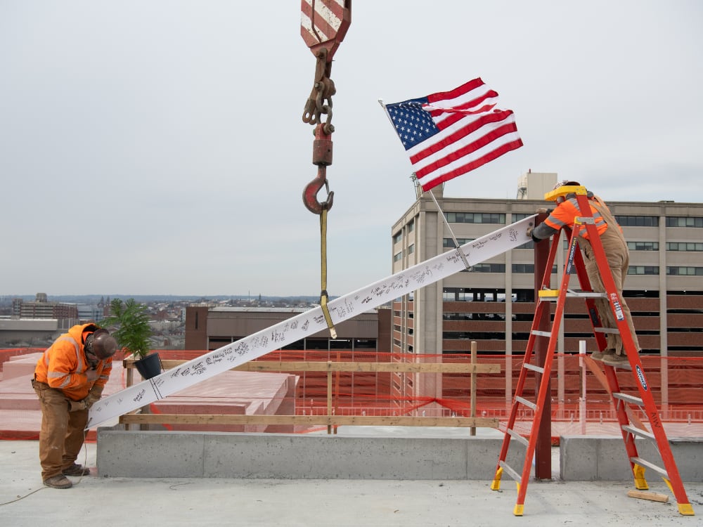 Two construction workers secure a signed steel beam at a rooftop site with an American flag waving nearby.