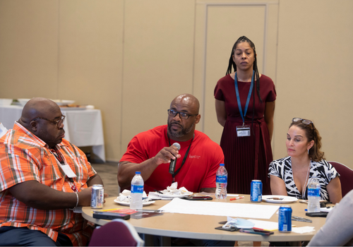 Tyrone Roper, holding microphone, provides input during a strategic diversity planning session while tablemates Hershaw Davis Jr. and Nicole Palmore, and Reetta Gach (standing) listen.