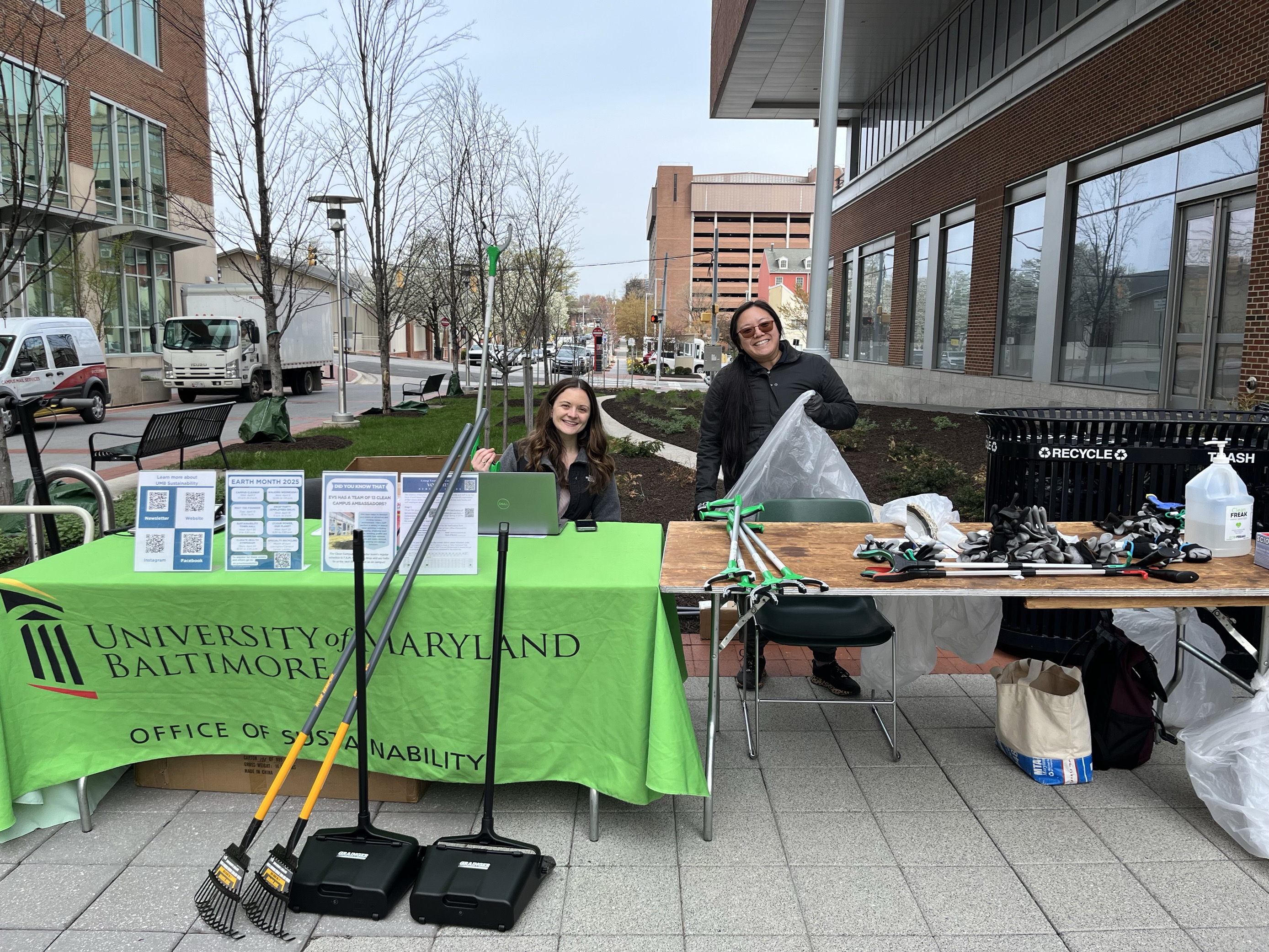 Two sustainability office members standing behind a table outside. The table has cleanup equipment and one of the members is holding a trash can